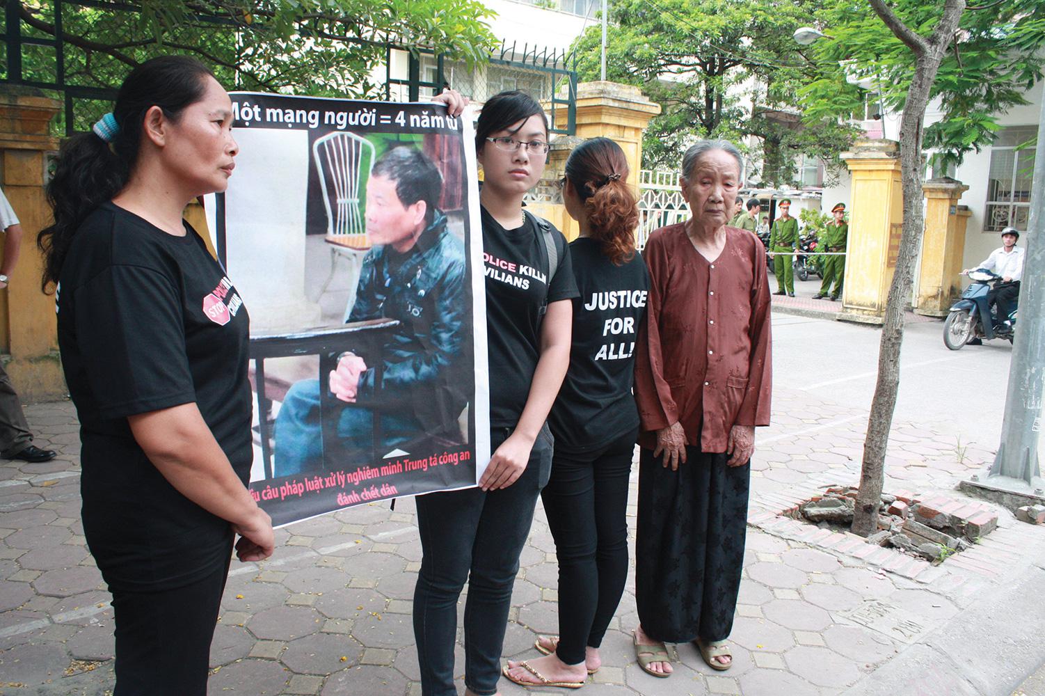 The family of Trinh Xuan Tung (his mother Nguyen Thi Cuc, his wife Nguyen Thi Mien, and his two daughters Trinh Kim Tien and Trinh Cam Tu) outside the court on July 17, 2012, waiting to attend appellate proceedings in the trial of former Col. Nguyen Van Ninh, convicted of causing Trinh Xuan Tung’s death. The poster features a picture of the perpetrator and slogans calling on authorities to impose a longer prison term. © 2012 Dan Lam Bao & contributors.