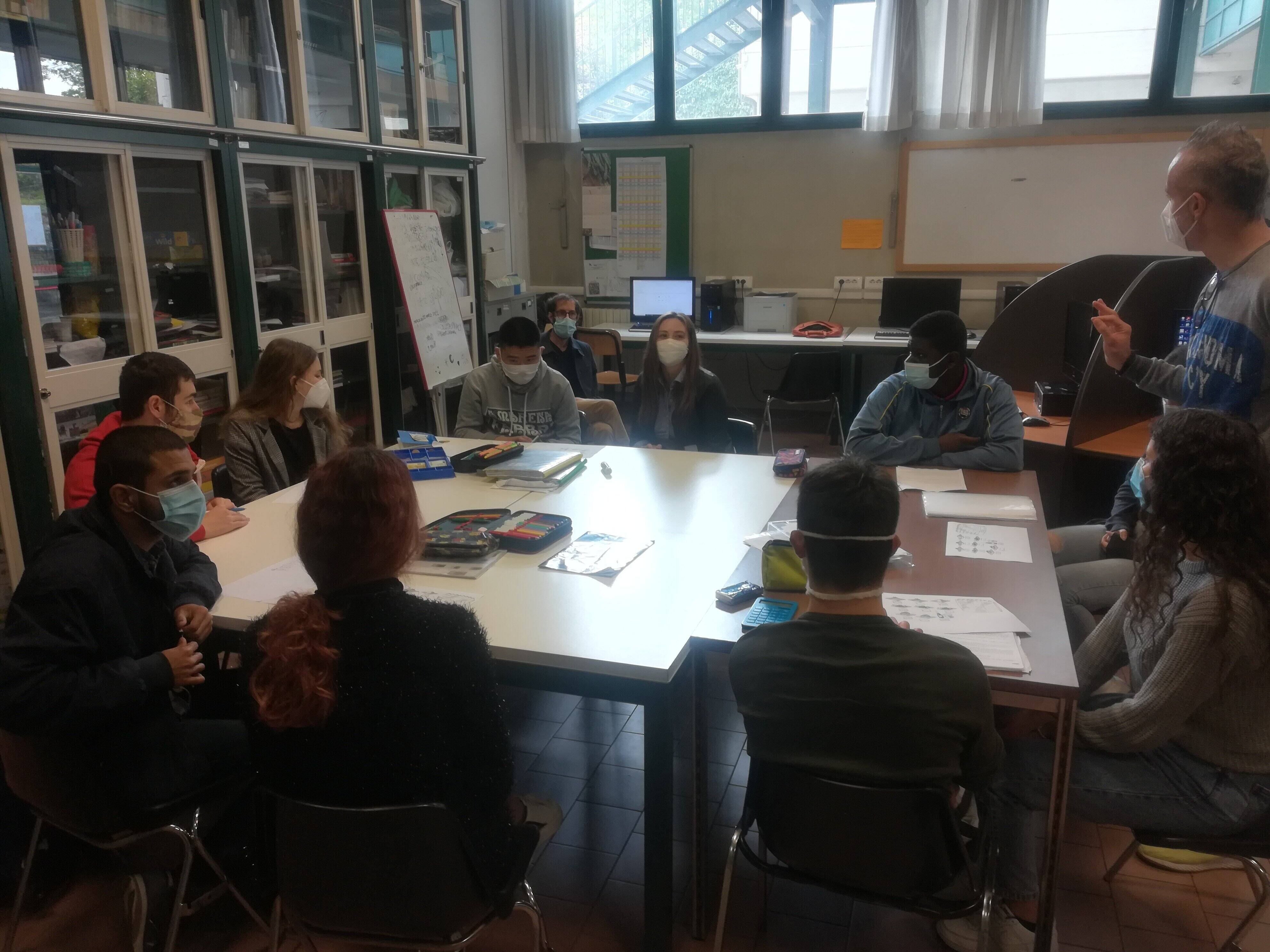 Studenti seduti intorno a una tavola, mentre ascoltano un insegnante.