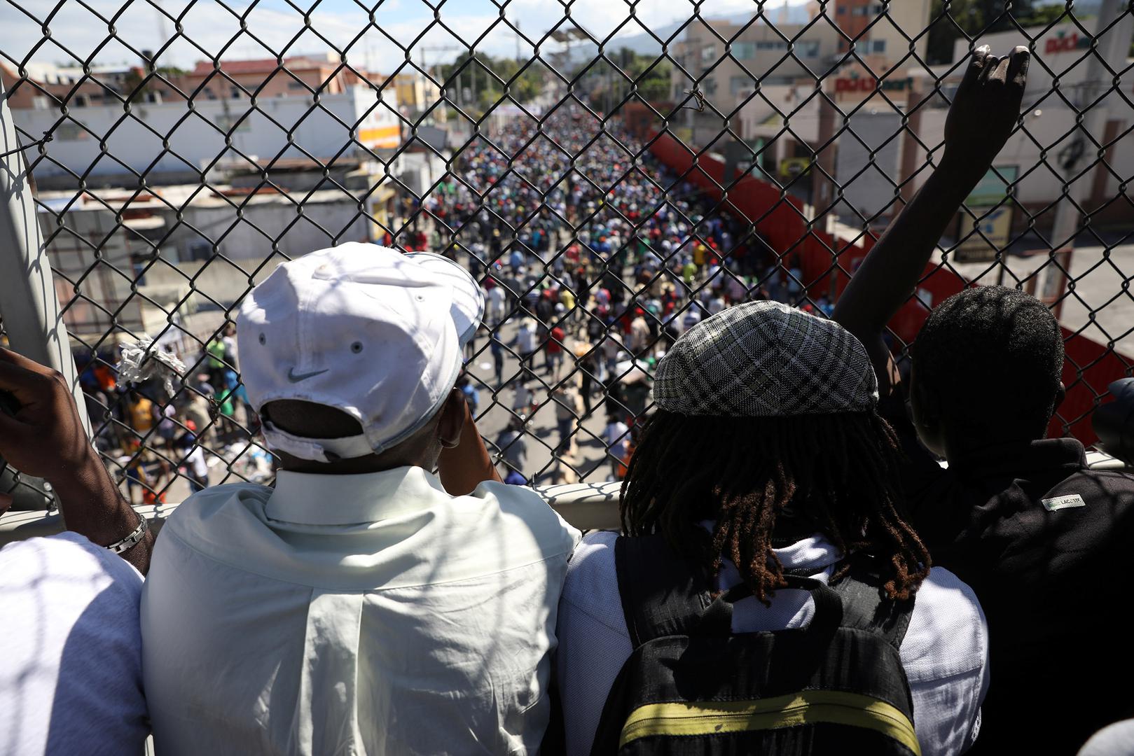 Men look at protesters marching to demand an investigation into what they say is the alleged misuse of Venezuela-sponsored PetroCaribe funds, in Port-au-Prince, Haiti, November 18, 2018. 