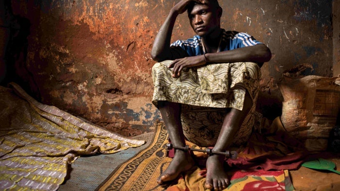 A man sitting on the floor in a dilapidated room, with his ankles locked by a metal bar.