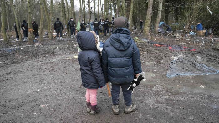 Two children wearing winter coats stand in a forest in front of a group of police officers