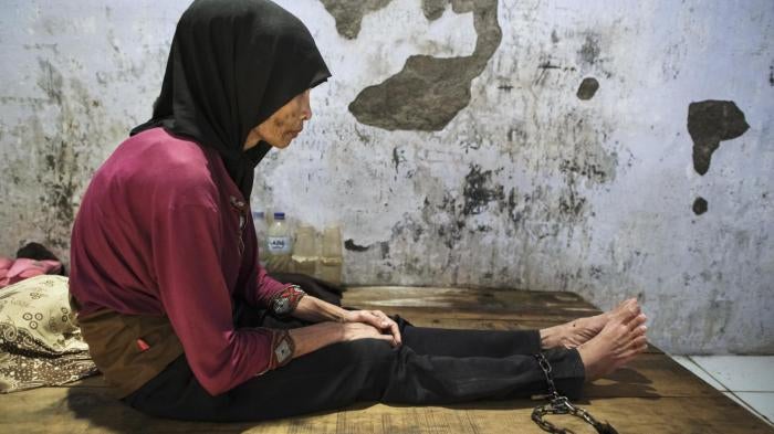 A woman with a scarf sits on a wooden platform to which she is shackled. 