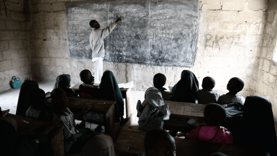 A picture of a teacher writing on a black board while students pay attention.