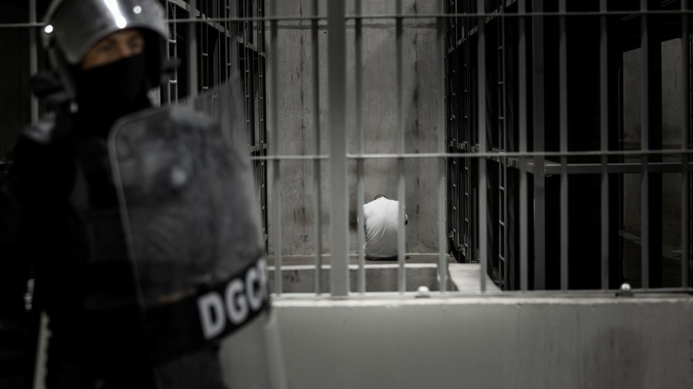 A person sits inside a cell while a guard with a riot shield stands outside