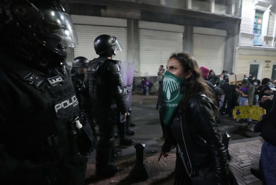 A woman wearing a green bandana over her face stands in front of a police officer with riot gear