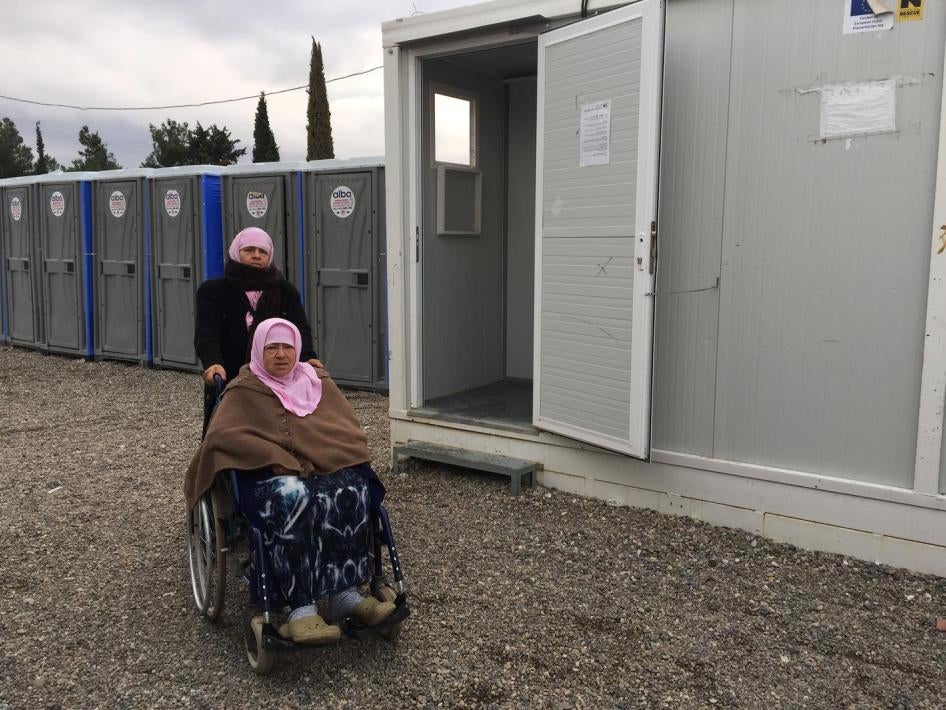 Naima, 70, an older woman with a disability from Aleppo, Syria, with her daughter Hasne, in front of the shower area in Cherso camp, Thessaloniki. The showers are not accessible for people who use a wheelchair.