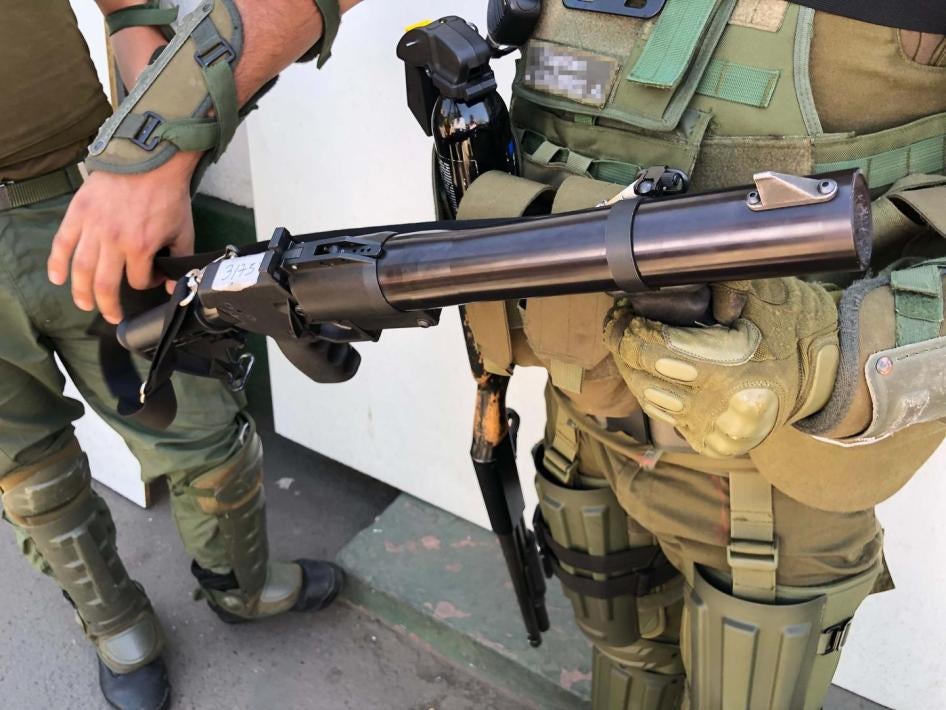 A police captain holds a teargas gun. A pellet shotgun hangs on his right side. 