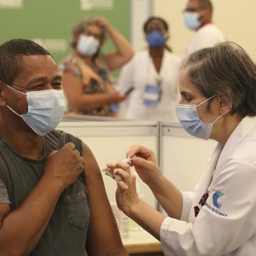 A white female healthcare professional wearing a mask, applying the covid-19 vaccine to a black male healthcare professional, wearing a mask, who is smiling