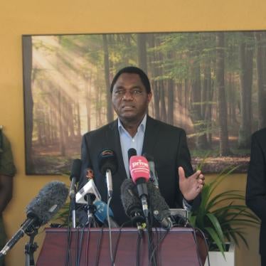 A man stands at a podium flanked by two people in face masks